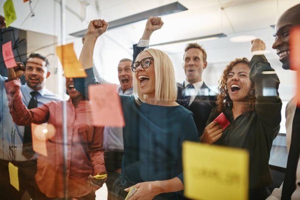 Diverse group of businesspeople cheering and fist pumping while brainstorming together with sticky notes on a glass wall in an office